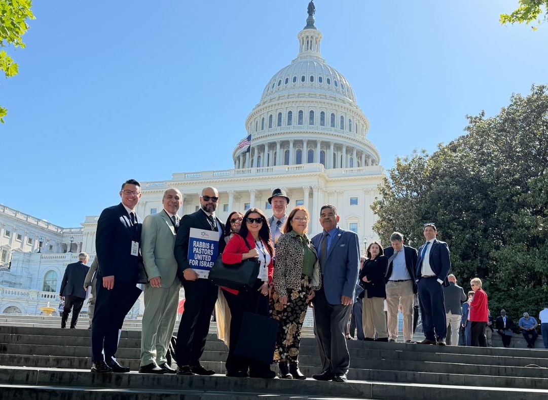 Group of professionals posing on the steps of the U.S. Capitol on a sunny day, with one person holding a sign reading 'Rabbis & Pastors United for Israel'.