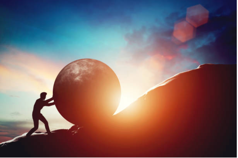 Silhouette of a person pushing a giant ball up a hill at sunset, with warm orange light behind them.
