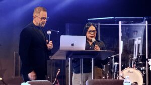 Two presenters stand at a lectern on a dimly lit stage, each with a microphone, a laptop on the podium, and blue lighting in the background.