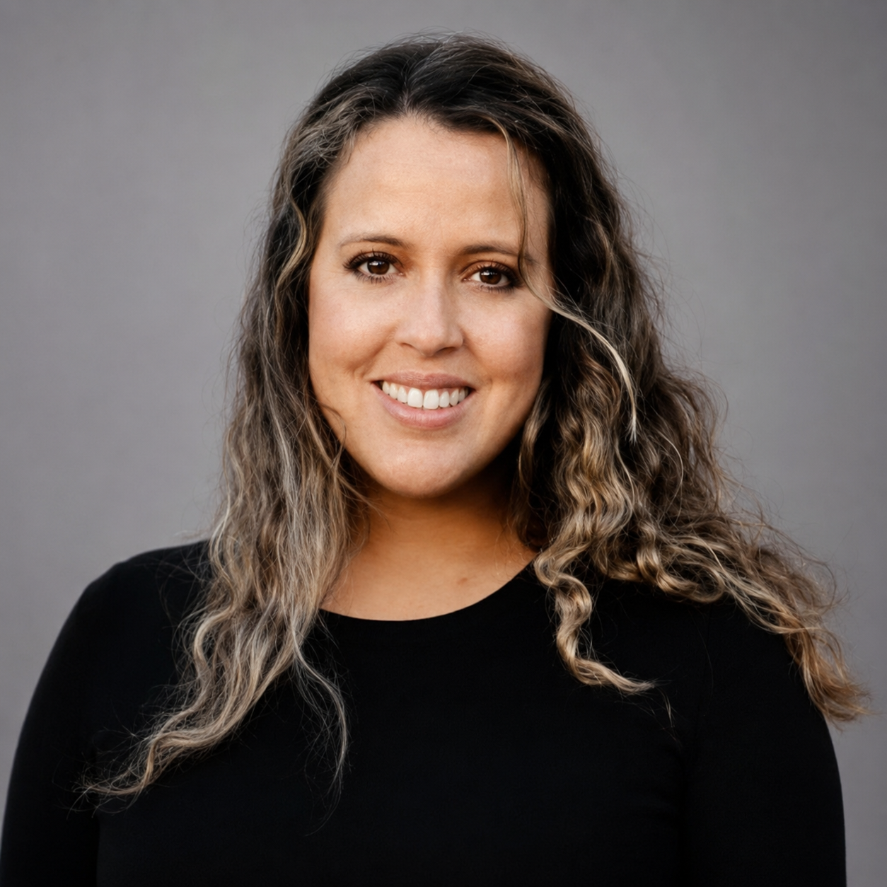 Portrait of a smiling woman with long wavy brown hair wearing a black top, against a neutral gray studio backdrop.