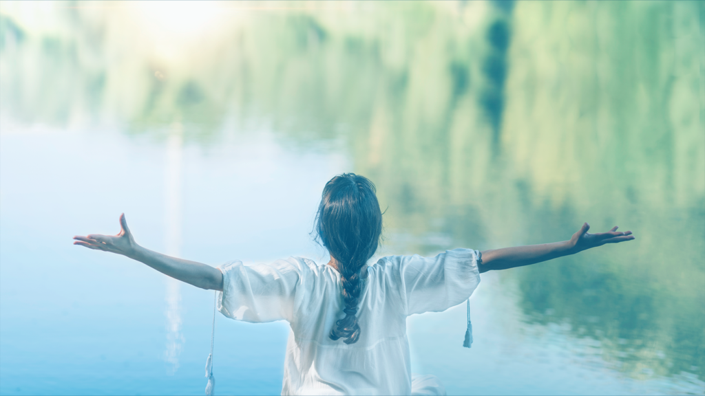 Person with braided hair wearing a white blouse stands with arms outstretched beside a calm lake, surrounded by trees.