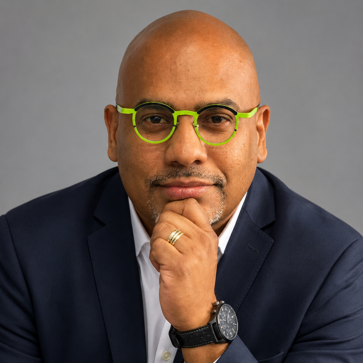 Professional head-and-shoulders portrait of a bald man in a navy suit with neon green round glasses, resting his chin on his hands.