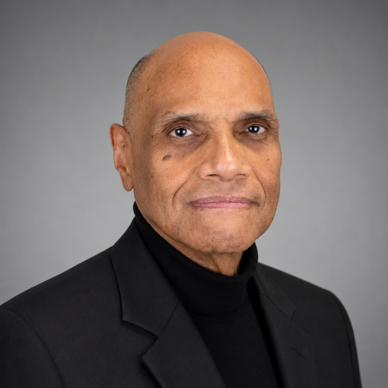 Professional headshot: bald man in a black blazer and turtleneck, facing the camera against a gray backdrop.
