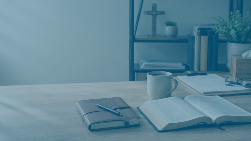 Open hardcover book on a wooden desk with a pen-tied notebook, a coffee mug, and reading glasses nearby; bookshelf in background.