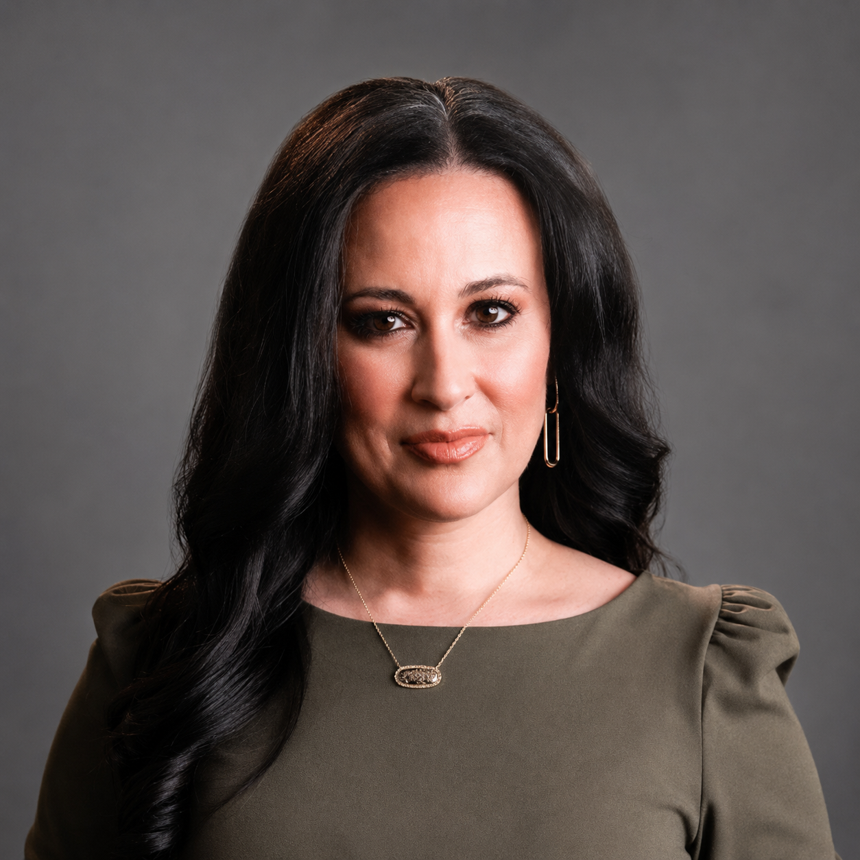 Professional head-and-shoulders portrait of a woman with long dark hair, olive blouse, and gold jewelry against a gray backdrop.