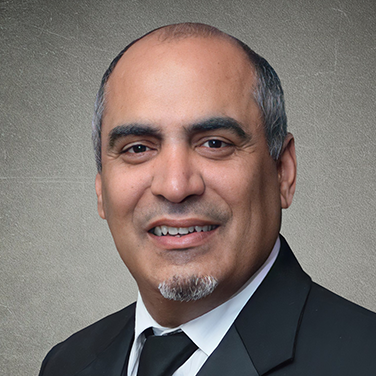 Professional headshot: smiling man in a black suit and white shirt against a neutral backdrop.