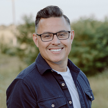 Smiling man with short dark hair and glasses, wearing a navy button-down over a white tee, outdoors with greenery in the background.