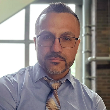 Professional headshot of a man with short hair and glasses, wearing a light blue dress shirt and patterned tie in an office setting