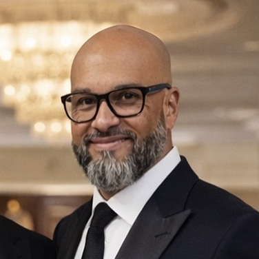 Bald man with glasses and a trimmed gray beard, wearing a dark suit and tie at a formal event, smiling.