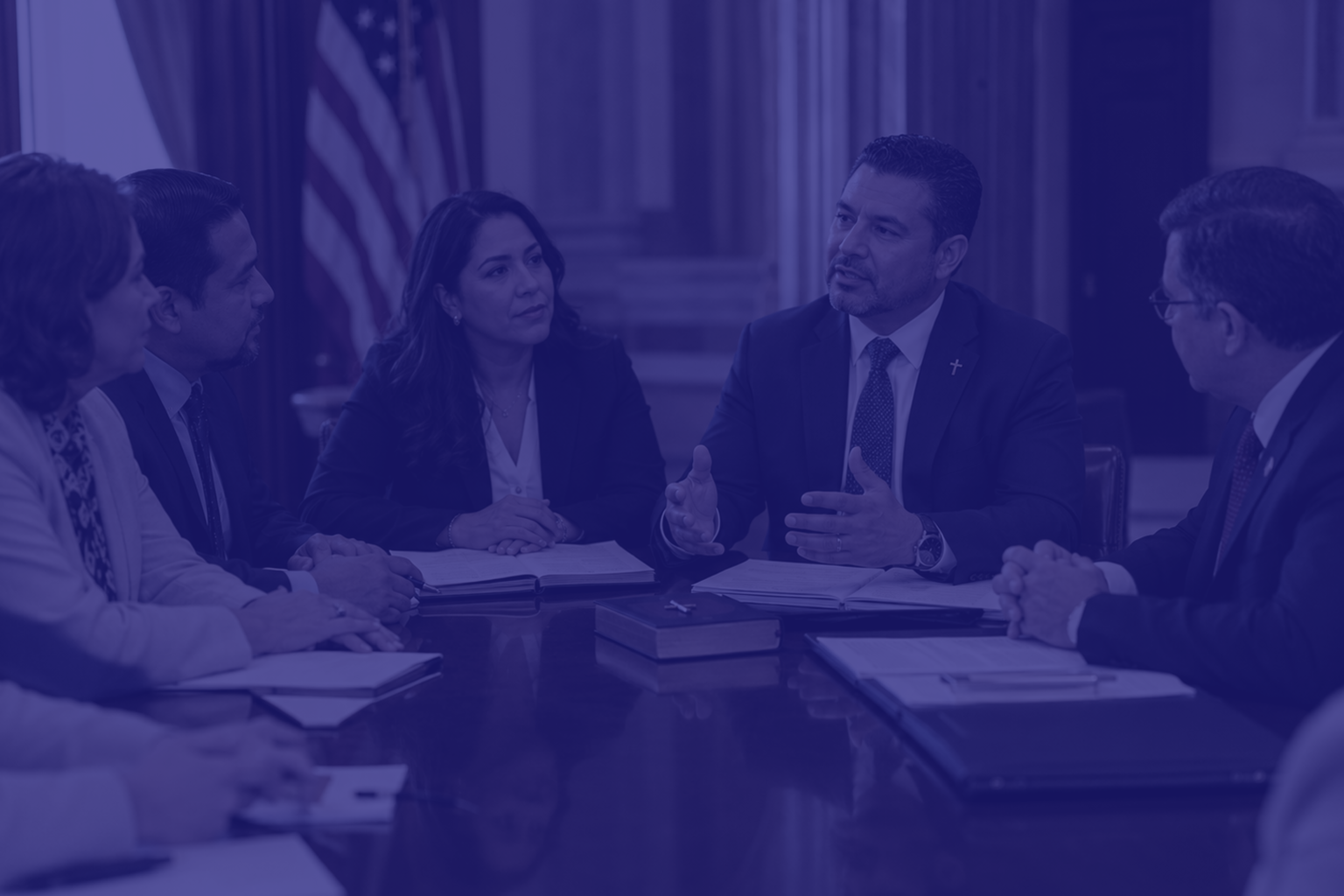 Five professionals around a conference table in a formal meeting; a man in a suit speaks with hand gestures while others listen and take notes.