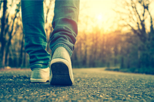 Person walking away on a sunlit forest path, close-up of jeans and sneakers from behind.
