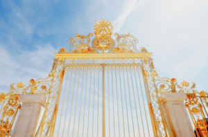 Golden ornate gate with intricate scrollwork and crest, set between stone pillars against a blue sky.