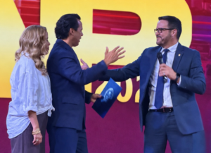 Three adults on stage greet each other: a man in a blue suit hands a blue folder to another man holding a microphone, while a woman watches beside them.