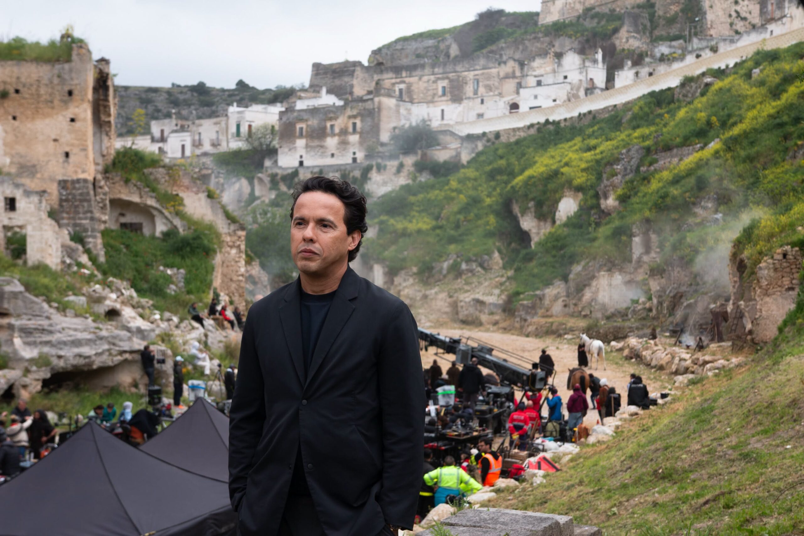 Man in a black suit stands in front of a hillside film set with ruins, crew, and equipment in the background.