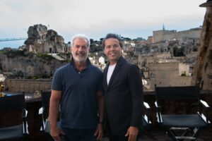 Two men smiling at the camera on a terrace with ancient stone ruins and a town behind them.