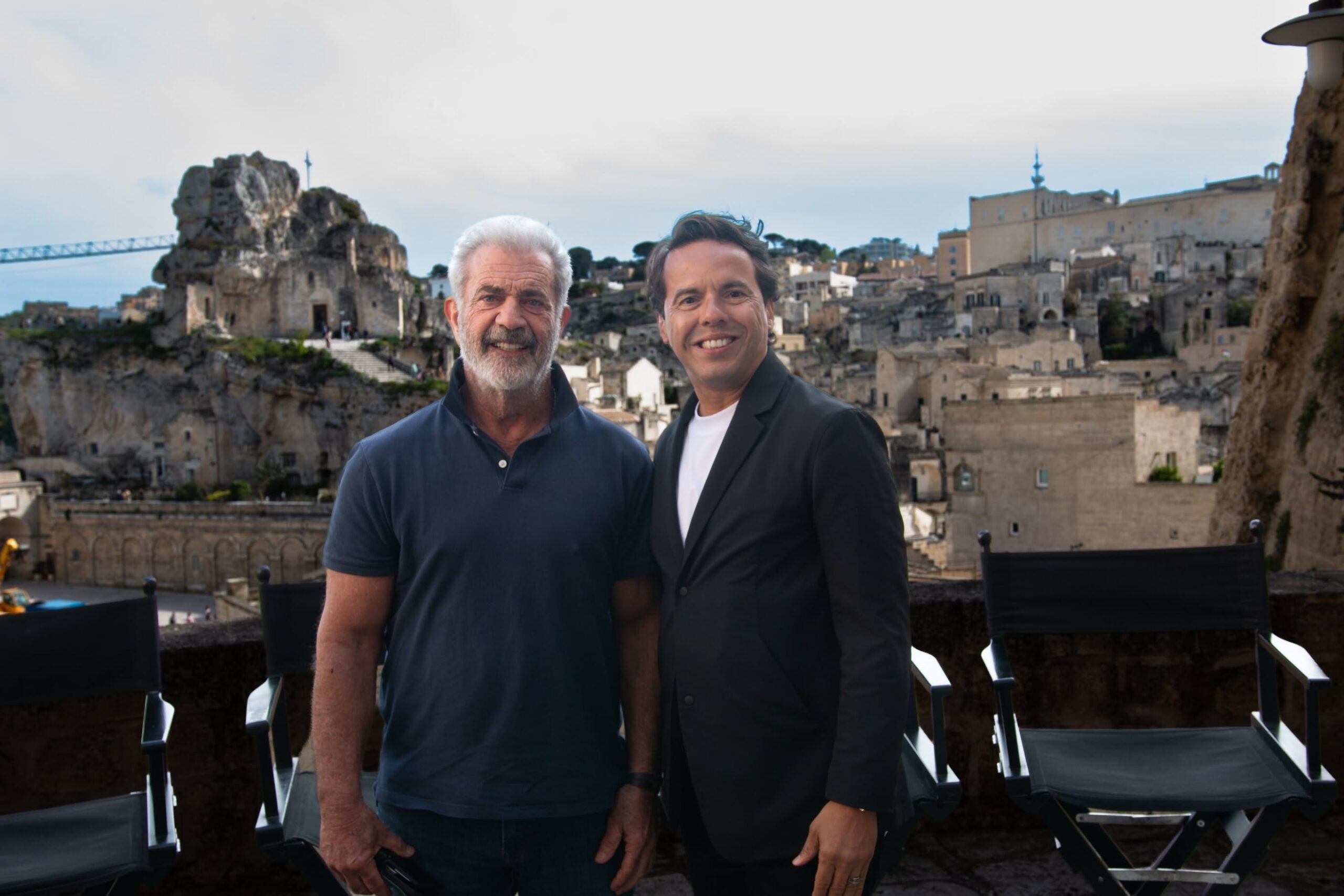 Two men smiling at the camera on a terrace with ancient stone ruins and a town behind them.