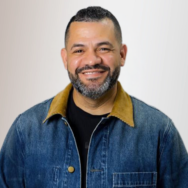 Portrait of a smiling man in a denim jacket against a light neutral background.