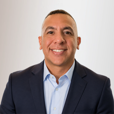 Professional headshot: smiling man in a navy blazer and light blue shirt against a light background on a neutral backdrop.