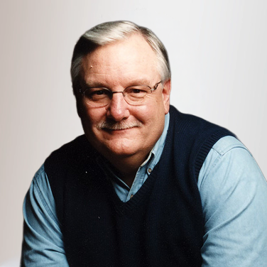 Portrait of an older man with gray hair and glasses, in a light blue shirt and navy sweater vest, smiling at the camera.