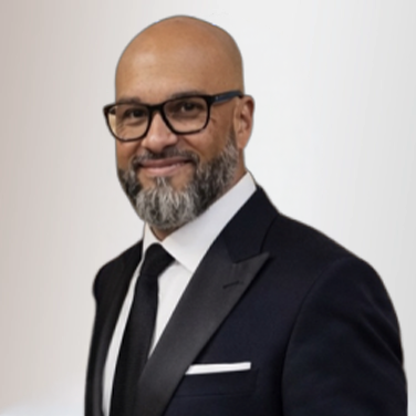 Portrait of a bald man with glasses and a gray beard, wearing a black suit and tie with a white pocket square, facing slightly to the right.