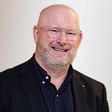 Smiling middle-aged man with glasses and a short gray beard, wearing a black blazer and dark shirt, against a light background.