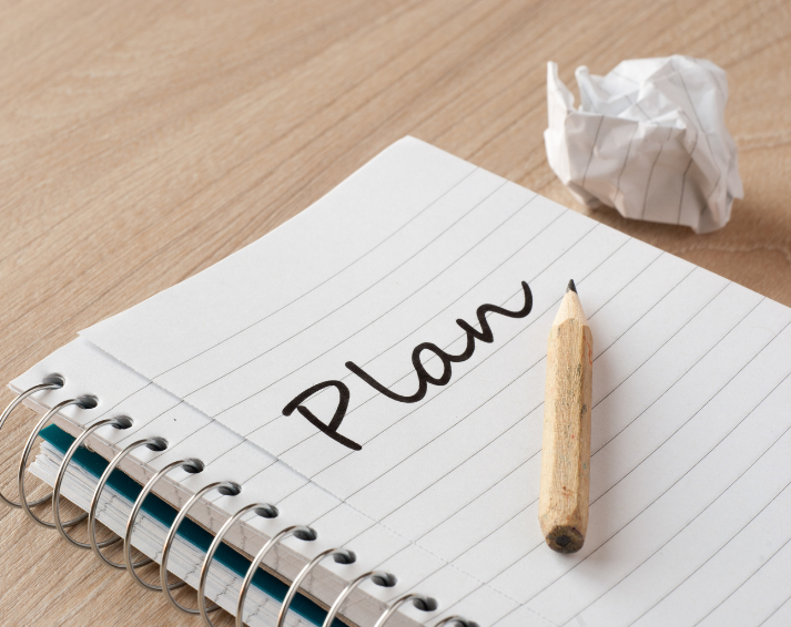 Notebook page with the word 'Plan' written in ink, a wooden pencil resting on the lines, and a crumpled sheet of paper on a wooden desk.