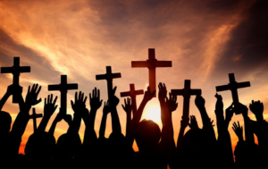 Silhouetted crowd raises wooden crosses toward a sunset sky, conveying a religious gathering or prayer scene.