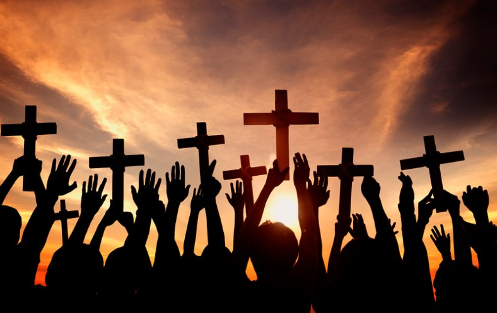 Silhouetted crowd raises wooden crosses toward a sunset sky, conveying a religious gathering or prayer scene.