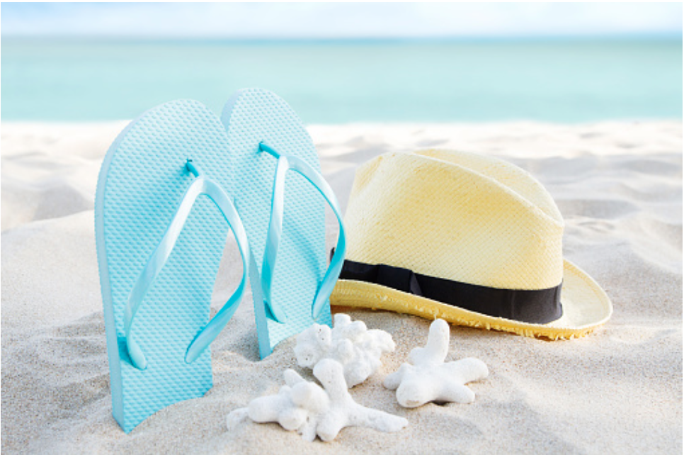Blue flip-flops and a straw hat resting on light sand at the beach with white coral nearby and turquoise sea in the background.